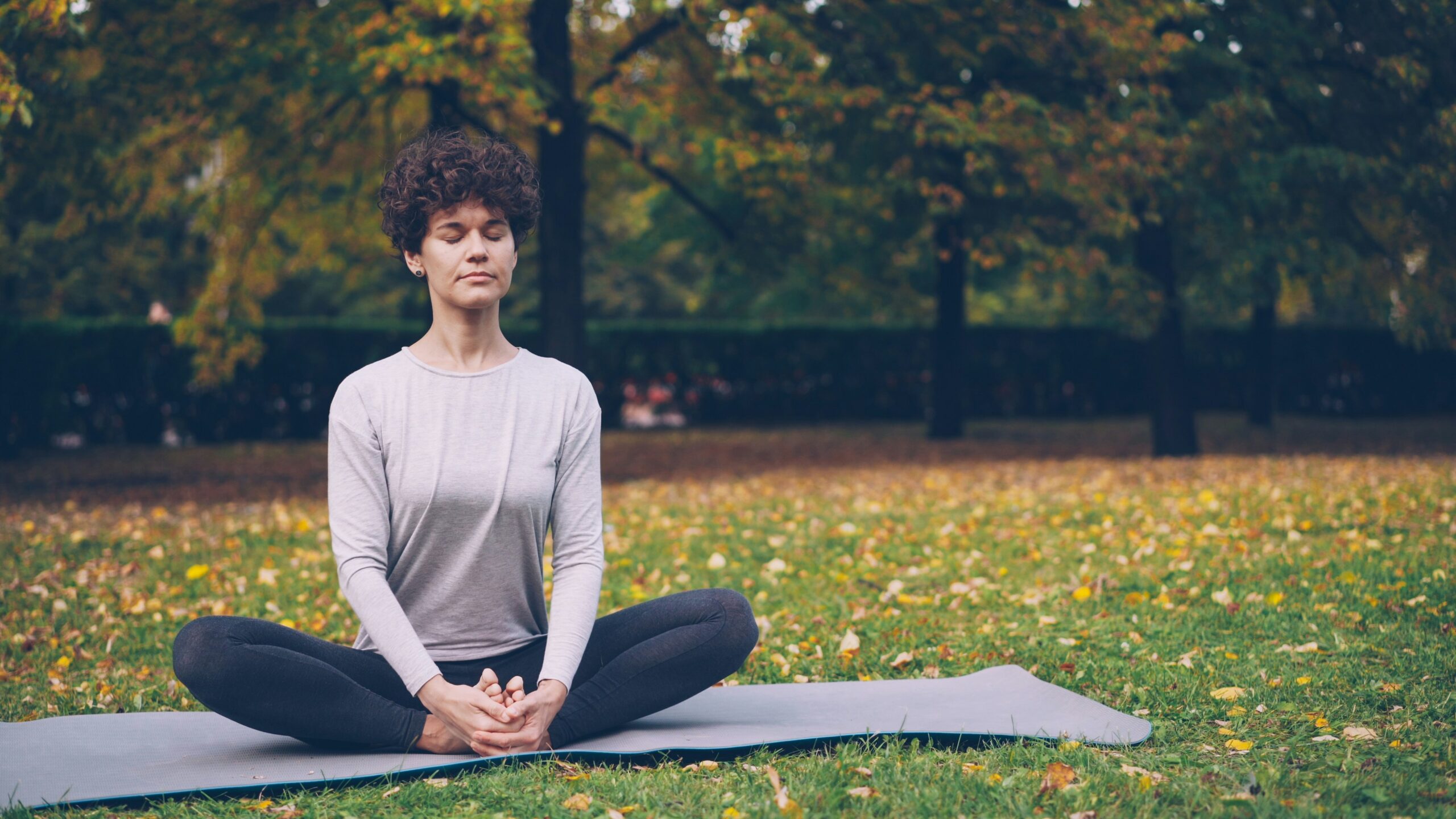 Flexible young lady is sitting on mat in yoga pose enjoying fresh air, peace and relaxation. Healthy lifestyle for urban people, active youth and autumn nature concept.